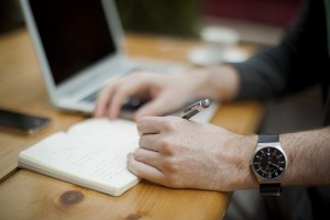 Man writing in a notebook; computer in the background.